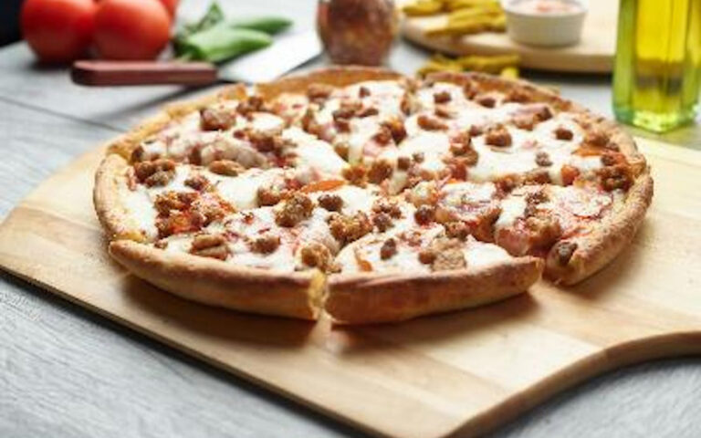 A freshly baked pizza with sausage and melted cheese sits on a wooden pizza peel. In the background, tomatoes, green leaves, and dipping sauce are slightly out of focus.