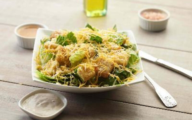 The Spring Caesar Chicken Salad is served in a square white bowl, topped with grated cheese and croutons, accompanied by small bowls of dressing and seasonings on a light wooden table with a fork and knife beside it.