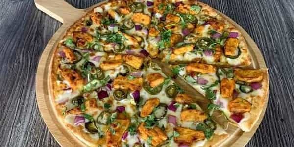 Achari Chicken Pizza on a wooden board, topped with chicken, jalapeños, and red onions.