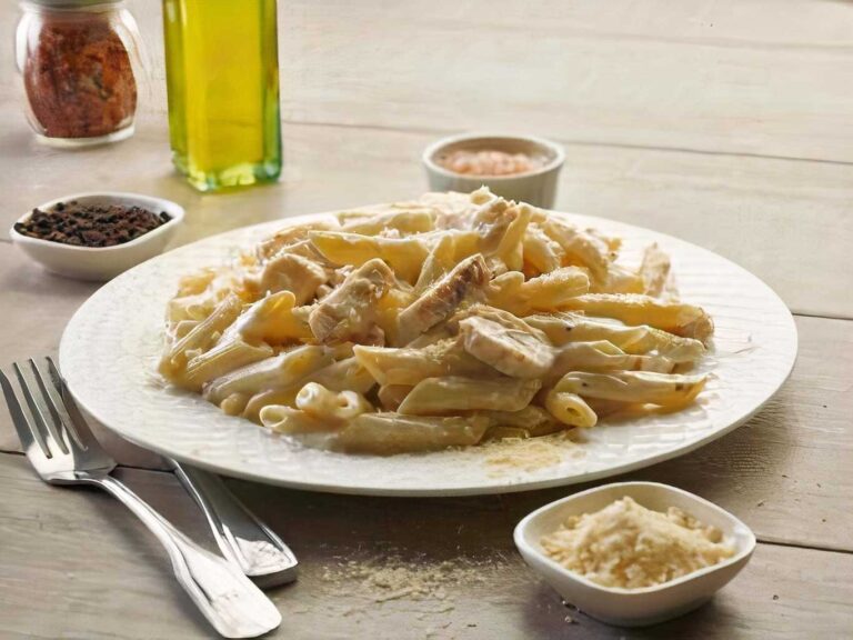 Alfredo chicken pasta on a white plate, with parmesan cheese and spices on a wooden table.