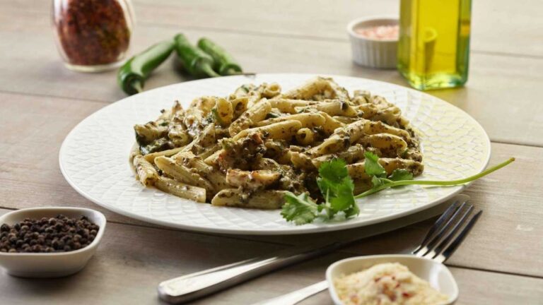 Pesto chicken pasta on a white plate, garnished with cilantro. Peppers and spices surround the dish.
