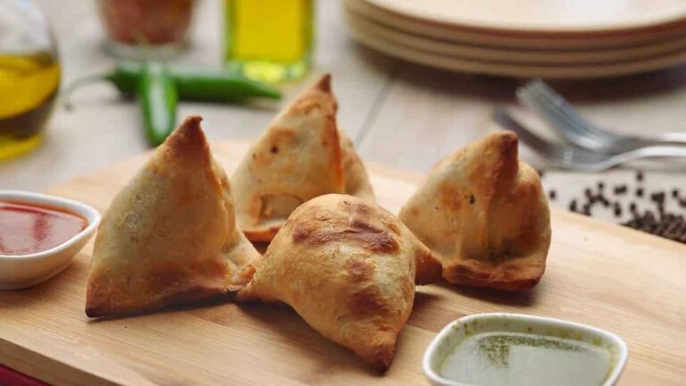 Four golden samosas on a wooden board with dipping sauces. Samosa (4 pcs) displayed.